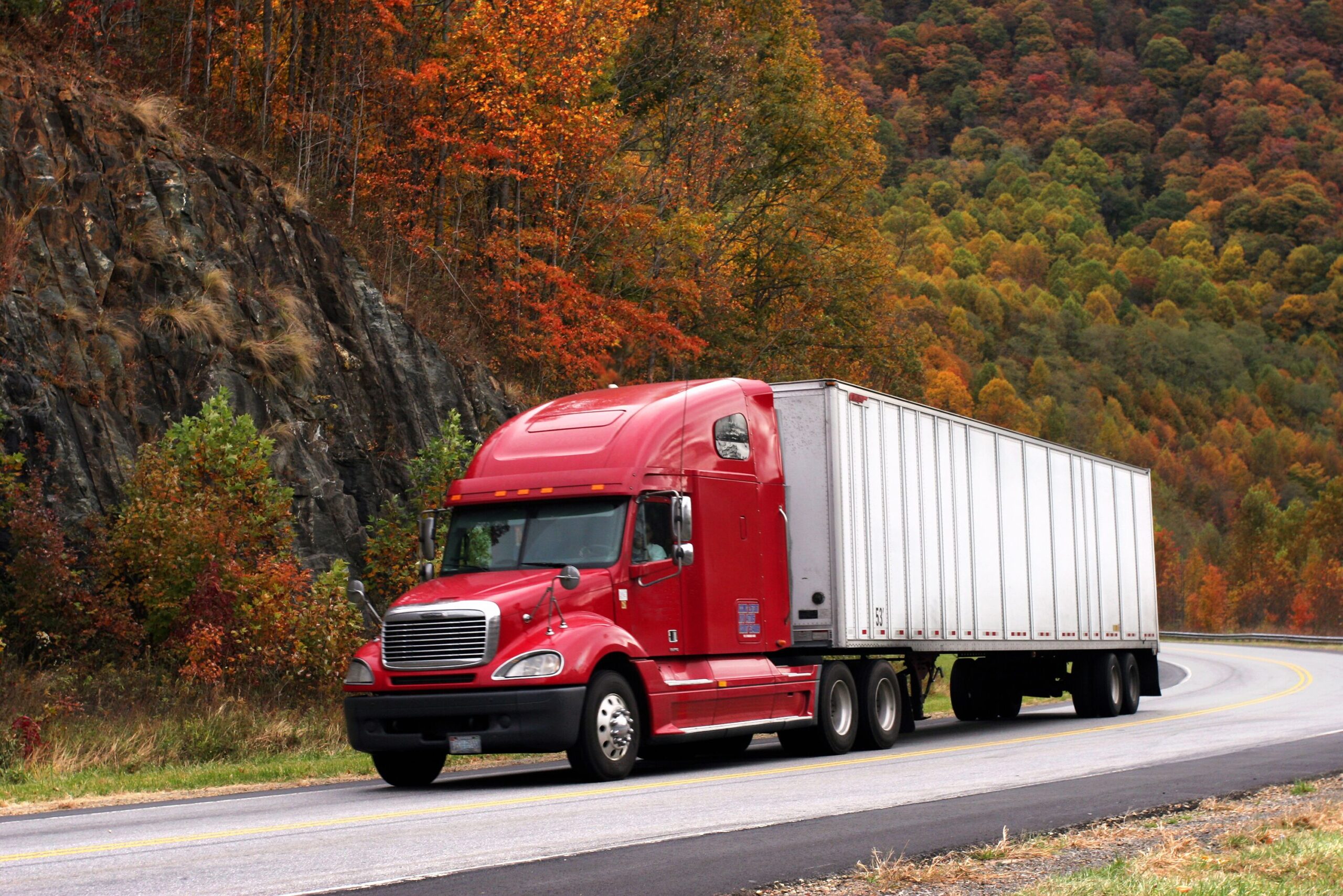 A red semi-truck drives along a winding road, surrounded by vibrant autumn foliage and rocky terrain, highlighting the beauty of fall travel.