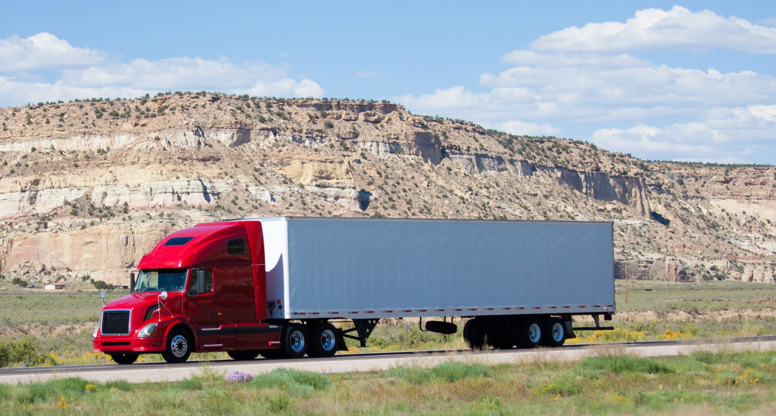 Red semi-truck on a highway against a backdrop of rocky cliffs and clear blue skies, highlighting transport in a scenic landscape.