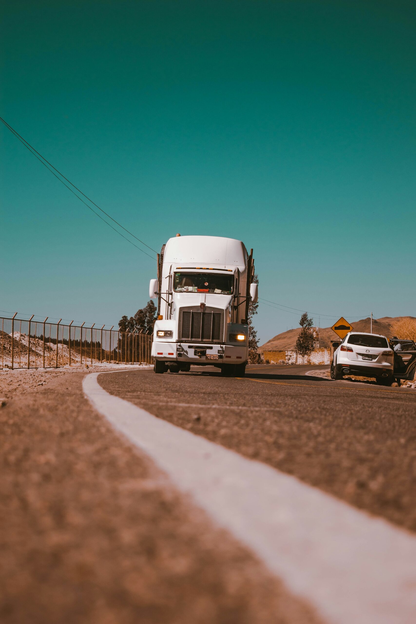 A large white truck drives along a winding road, flanked by a fence and a parked car, under a clear blue sky.