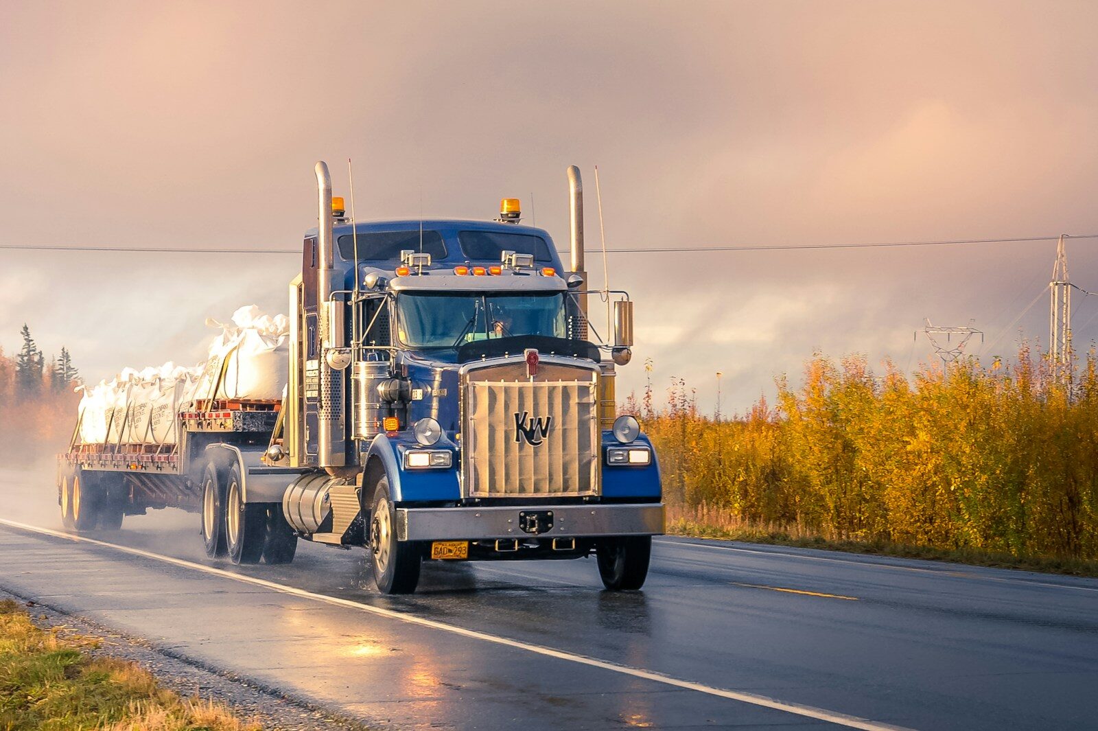 A blue semi-truck transporting white wrapped bales drives along a wet road amid an autumn landscape, showcasing freight transportation in action.