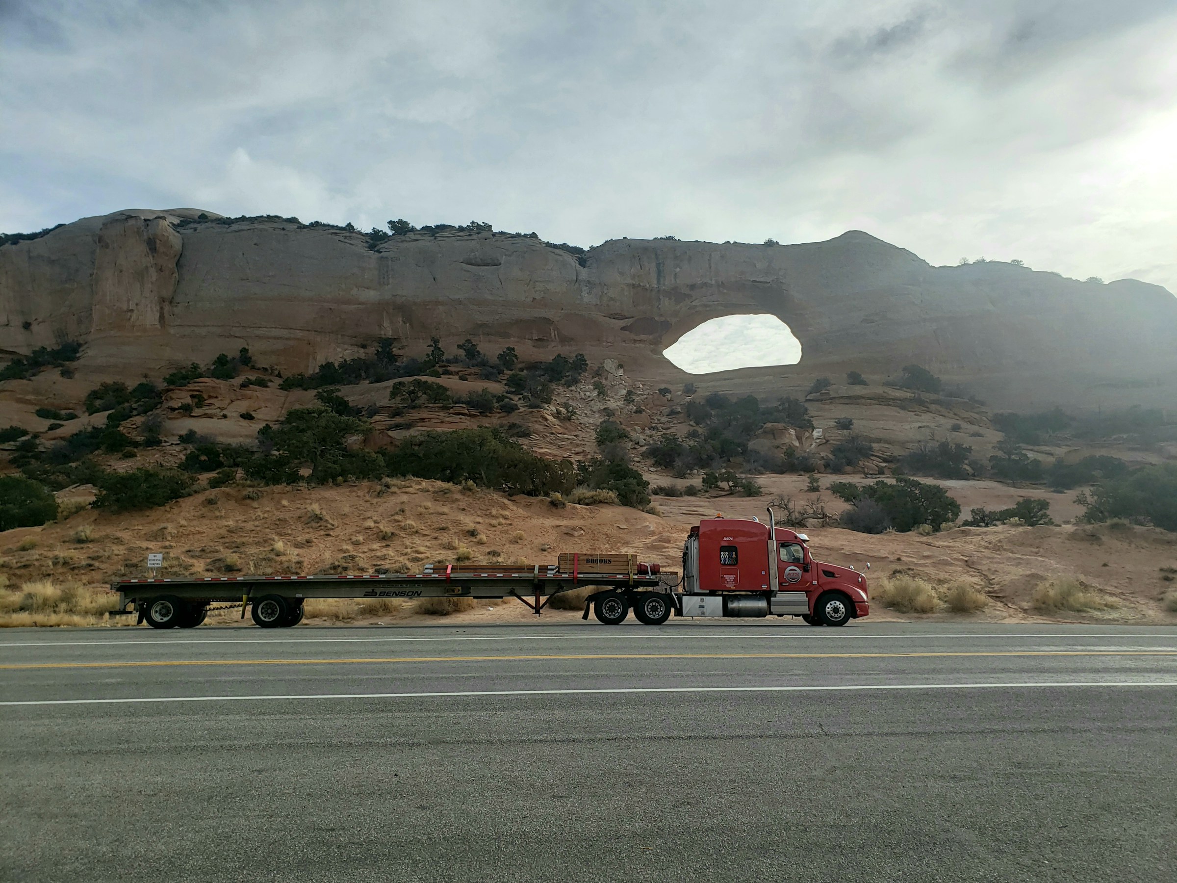 Red truck parked beside a scenic rock formation featuring a large natural arch. The landscape showcases unique geological features and vegetation.